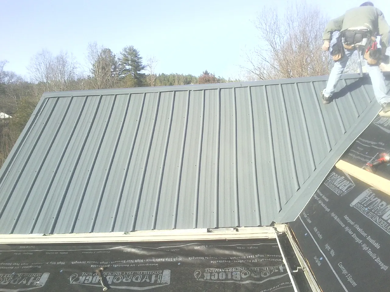Crew installing dark green standing seam panels over synthetic underlayment in East Freedom