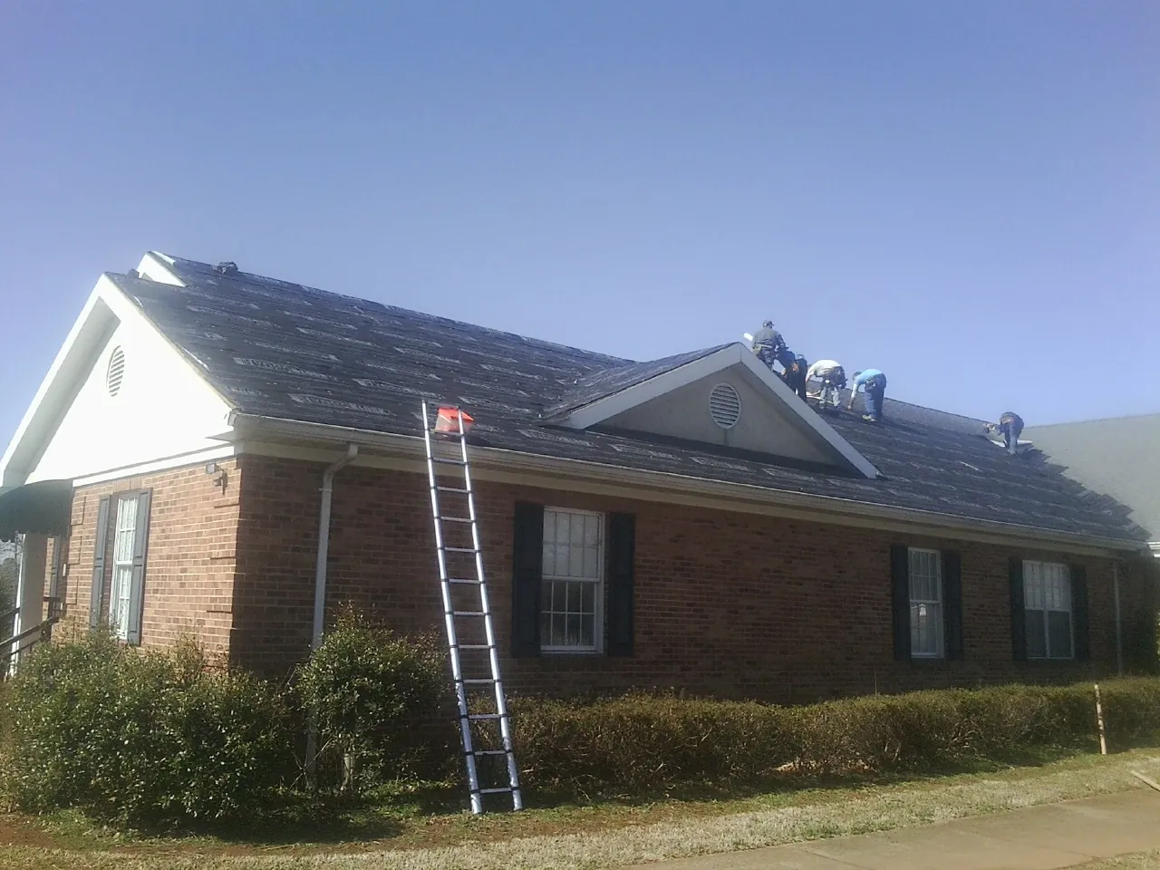Crew tearing shingles off a brick ranch roof with a ladder set — Metal Roof Repair work by our crew in East Freedom