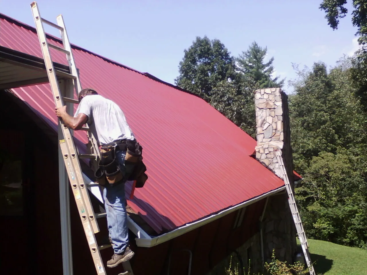 Roofer on a ladder beside a red metal roof and stone chimney — Soffit & Fascia Repair by Roofing Brothers in East Freedom